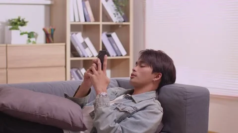 Sitting on the sofa in the house, an Asian man works on his smartphone.Man wo Stock Footage 172425793