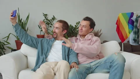 Sitting on the sofa, two handsome lgbt guys are taking funny photos, laughing Stock Footage 174479125
