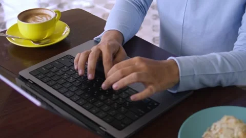 Sitting at the table with a cup of coffee, typing on laptop keyboard Видео 267185202