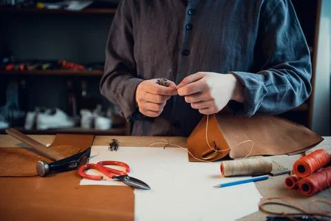 Sitting at a table in the workshop a young guy manually sews leather elements to Stock Photos