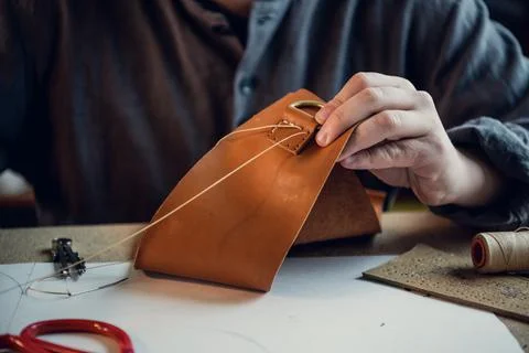 Sitting at a table in the workshop a young guy manually sews leather elements to Stock Photos