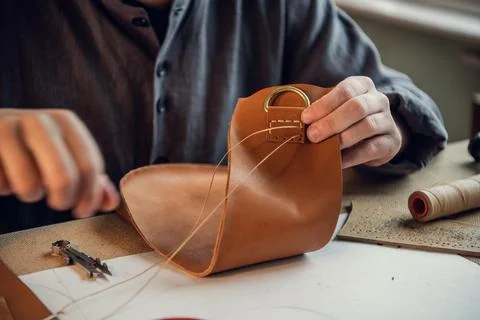 Sitting at a table in the workshop a young guy manually sews leather elements to Stock Photos