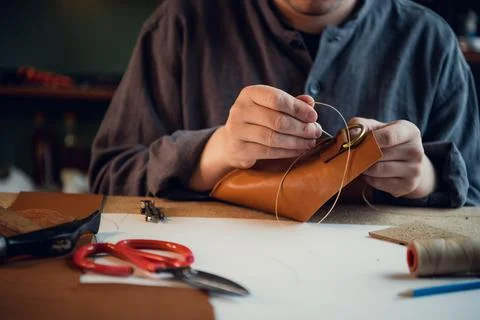 Sitting at a table in the workshop a young guy manually sews leather elements to Stock Photos