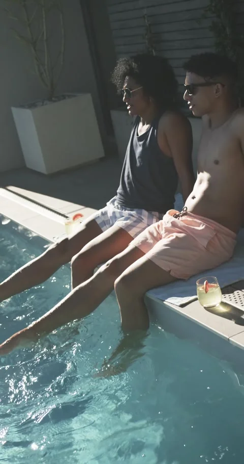 Sitting two men dipping feet at pool patio, wearing sunglasses, peach shorts, Stock Footage 345218939