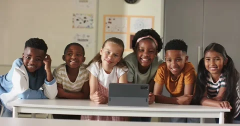 Six diverse schoolchildren leaning on table in classroom, peering at tablet on Stock Footage 338325882