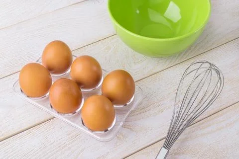Six raw chicken eggs in a transparent plastic tray, green bowl and whisk Stock Photos