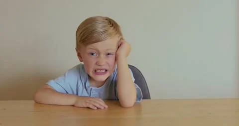 A six-year-old blond boy sits at a wooden table, showing an expression of anger. Vidéo 324818265