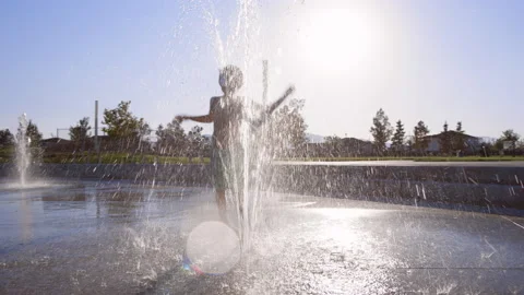 Six year old boy gleefully jumps in large sprinkler water jet at splash pad 스톡 동영상 233941837