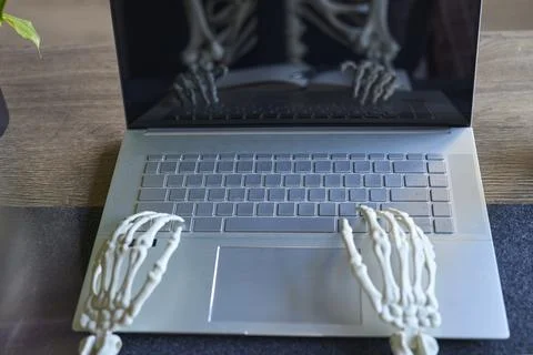 Skeleton hands positioned over laptop keyboard with reflection in dark screen Stock Photos