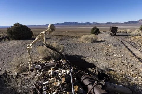 Skeleton using a rusty old wheelbarrow to load trash near tracks and an ore c Stock Photos