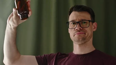 Skeptical man pours capsules from amber supplement bottle indoors during health Stock Footage 336623844