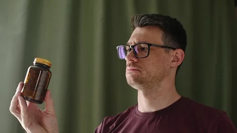 Skeptical man studies amber capsule bottle at home before careful medication and Stock Footage 336623837