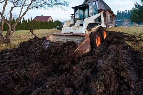 Skid steer loader churns mud by wood clad house and arborvitae Stock Photos