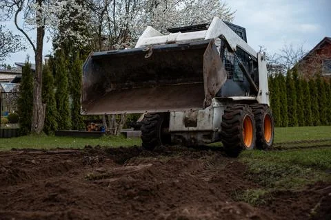 Skid Steer Loader on Fresh Soil Near Trees with White Blossoms Stock-Fotos