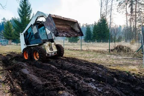 Skid Steer Loader Grading Muddy Path at Fenced Rural Worksite Stock Photos
