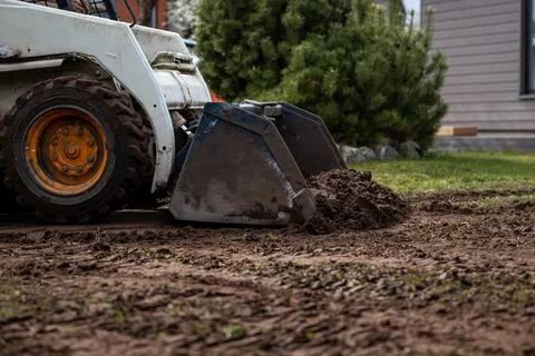 Skid Steer Loader Moving Soil Near Green Bush and Building Stock Photos