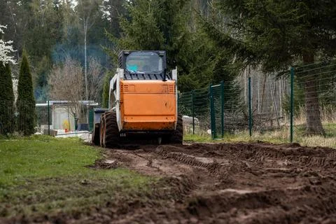 Skid Steer Loader Operating on Dirt Path Near Green Fence and Trees Stock Photos
