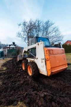 Skid steer loader in spring thaw mud near red roof house and trees Fotos Stock