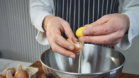 Skilled baker in striped apron carefully cracking eggs into mixing bowl Stock Footage 324575316