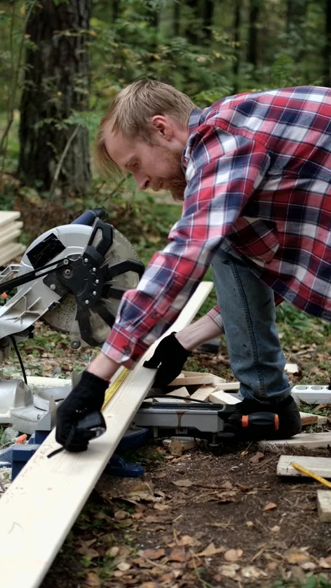 Skilled craftsman using a miter saw to cut wood in a forest setting, camera pans Stock Footage 322115255