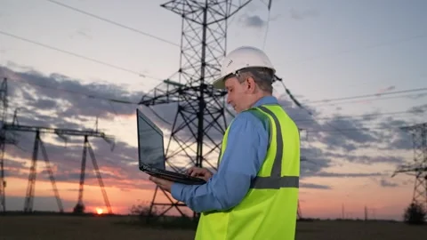 Skilled engineer works on laptop against power transmission lines at sunset Stock Footage 261438688