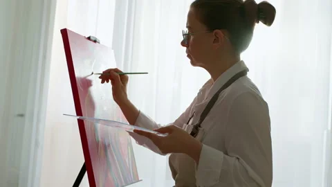 Skilled female painter creating expressive artwork on easel in bright studio.  Видео 331144877
