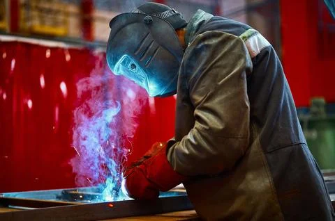 Skilled worker in protective mask welds metal part at plant Stock Photos