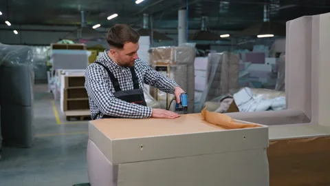 Skilled worker uses a pneumatic stapler to attach fabric to a sofa base frame Stock Footage 305612298