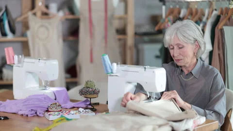 Skillful old tailoress sewing textile sitting at her workplace in tailor shop Stock Footage 342132499