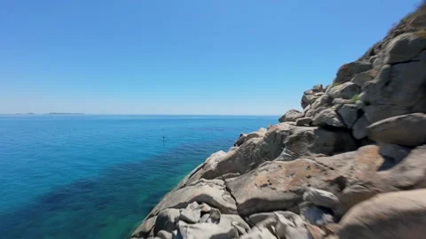 Skimming past sharp granite boulders and clear blue sea, an FPV drone flies low Stock Footage 342282104