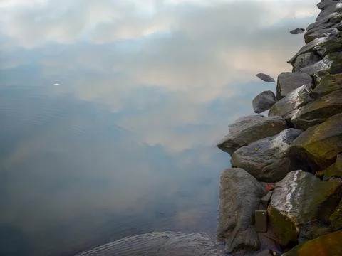 Sky Clouds Reflection on Calm Lake Water along Rocky Shoreline Stock Photos