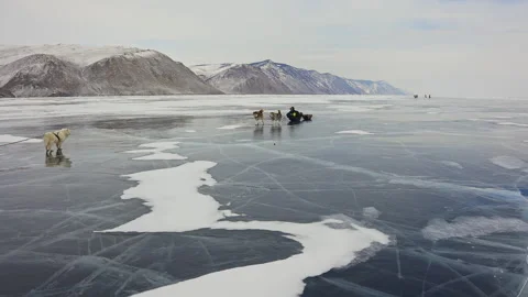 Sled dogs pulling sledges on cracked ice surface at frozen lake near snowy hills Stock Footage 165845531