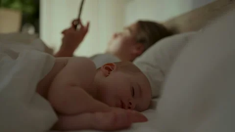 Sleeping baby resting on a bed while parent uses a smartphone in the background Vídeos de archivo 347412929