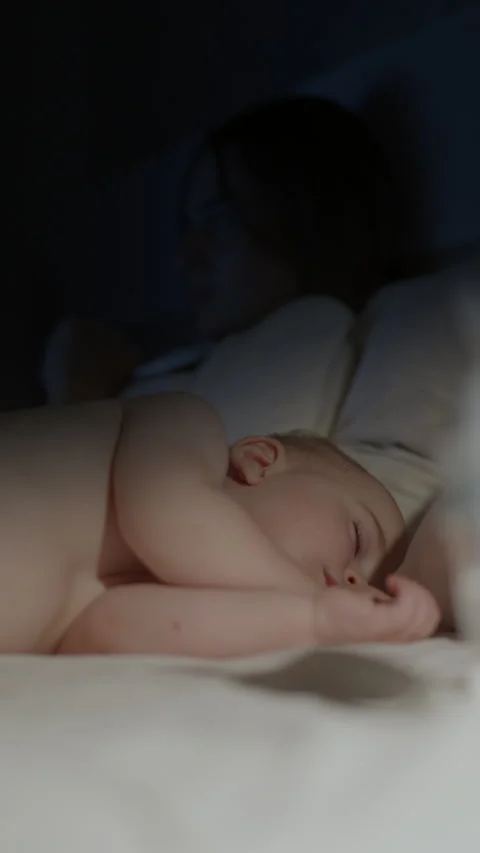 Sleeping infant peacefully rests on white bed sheets, with a blurred figure of a Stock Footage 341898698