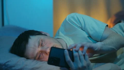 Sleepy man watching smartphone content lying in bed at night Видео 337892732