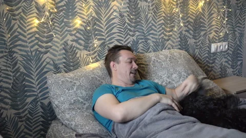 A sleepy middle-aged man plays with an elderly black dog while lying on a bed in 스톡 동영상 148446701