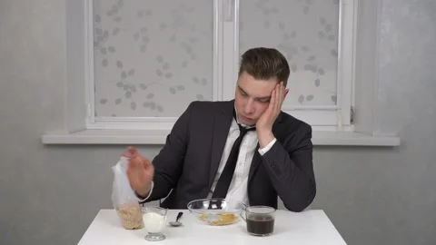 A sleepy young man in a suit eats cereal and drinks coffee. Young businessman at Stock Footage 150273986
