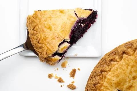 A slice of blueberry pie on a pie server being placed on a white square ceramic 스톡 사진