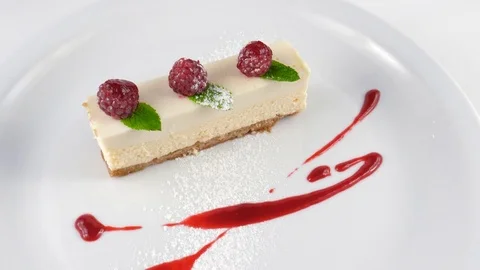 Slice of Cheesecake decorated with raspberry, icing sugar, mint leaf and red jam Stock Footage 79984360
