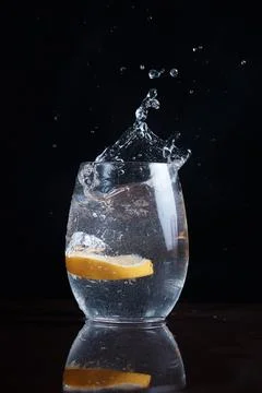 A slice of lemon falls into water in clear glass, splashes fly, citrus in glass Stock Photos