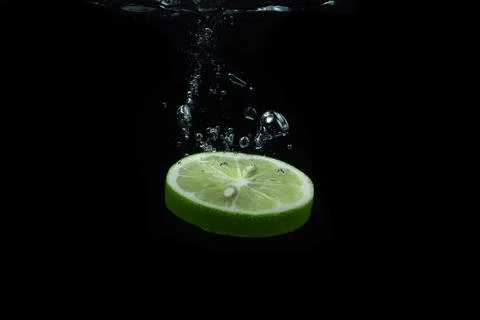 A slice of lime falls into the water on a black background. Sour fruit. Stock Photos
