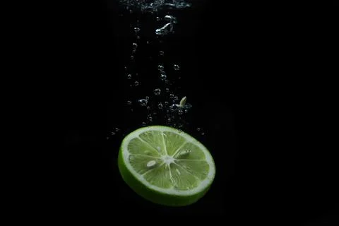 A slice of lime falls into the water on a black background. Sour fruit. Stock Photos