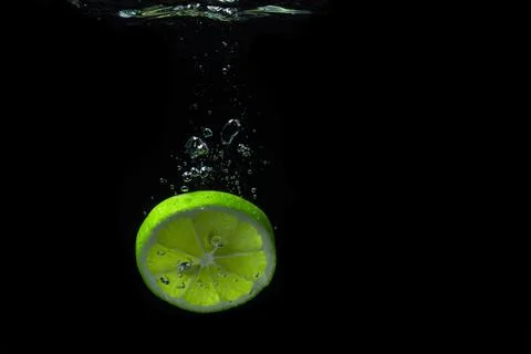 A slice of lime falls into the water on a black background. Sour fruit. Stock Photos
