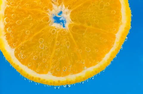 Slice of orange in the water with bubbles, on blue vibrant background Stock Photos