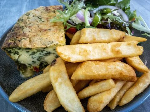 Slice of quiche on a serving plate placed beside some deep fried potato chips Stock Photos