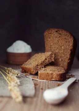 Sliced brown bread with ears of wheat, flour or salt over dark wooden backgro Stock Photos