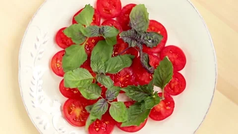 Sliced red cherry tomatoes and basil leaves on white plate rotating on wooden Video stock 157877605
