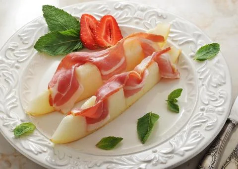 Slices of melon wrapped in bacon with mint leaves and strawberry on marble ba Stock Photos