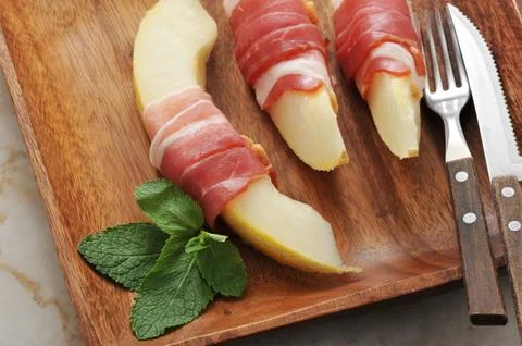 Slices of melon wrapped in bacon with mint leaves on a wooden platter Stock Photos