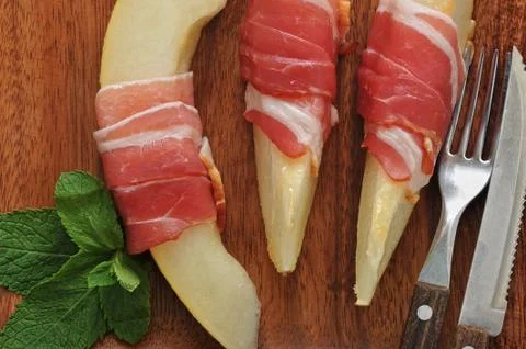 Slices of melon wrapped in bacon with mint leaves on a wooden platter Stock Photos